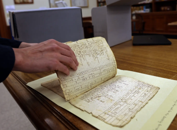 A researcher's hand flips the page of a frayed and fragile, historic document.